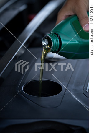 Person pouring fresh motor oil into a car engine during vehicle maintenance. Routine lubrication service performed in a workshop to protect engine components, reduce friction and support reliability. Person pouring fresh motor oil into a car engine during vehicle maintenance. Routine lubrication service performed in a workshop to protect engine components, reduce friction and support reliability. 142350513