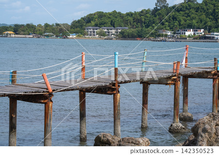Weathered wooden walkway with rope railing extending above rocky coast in Thailand, connecting local waterfront area while overlooking tropical sea. Traditional tropical maritime landscape. Weathered wooden walkway with rope railing extending above rocky coast in Thailand, connecting local waterfront area while overlooking tropical sea. Traditional tropical maritime landscape. 142350523