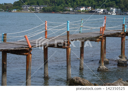 Weathered wooden walkway with rope railing extending above rocky coast in Thailand, connecting local waterfront area while overlooking tropical sea. Traditional tropical maritime landscape. Weathered wooden walkway with rope railing extending above rocky coast in Thailand, connecting local waterfront area while overlooking tropical sea. Traditional tropical maritime landscape. 142350525