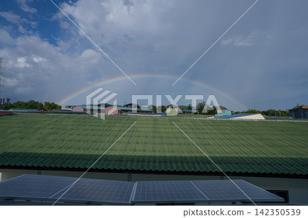 Full rainbow arching across sky above rooftops and photovoltaic solar panels, combining natural weather beauty with renewable energy and sustainable urban living. Renewable energy infrastructure. Full rainbow arching across sky above rooftops and photovoltaic solar panels, combining natural weather beauty with renewable energy and sustainable urban living. Renewable energy infrastructure. 142350539