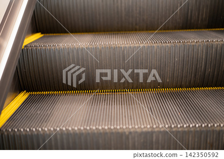 Empty escalator steps with yellow safety edges inside public building. Urban transportation infrastructure, pedestrian mobility, mechanical design and everyday travel. Public safety, accessibility. Empty escalator steps with yellow safety edges inside public building. Urban transportation infrastructure, pedestrian mobility, mechanical design and everyday travel. Public safety, accessibility. 142350592