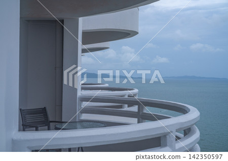 Seaside hotel balcony with curved white railing, chair and table overlooking blue ocean and island. Tropical travel, peaceful vacation lodging and relaxation. Coastal tourism. Private hotel balcony. Seaside hotel balcony with curved white railing, chair and table overlooking blue ocean and island. Tropical travel, peaceful vacation lodging and relaxation. Coastal tourism. Private hotel balcony. 142350597