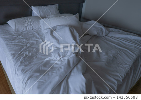 Unmade hotel bed with rumpled white sheets, pillows and duvet in dim guest room after sleep. Everyday travel accommodation, morning mess, comfort, rest and realistic hospitality lifestyle. Lodging. Unmade hotel bed with rumpled white sheets, pillows and duvet in dim guest room after sleep. Everyday travel accommodation, morning mess, comfort, rest and realistic hospitality lifestyle. Lodging. 142350598