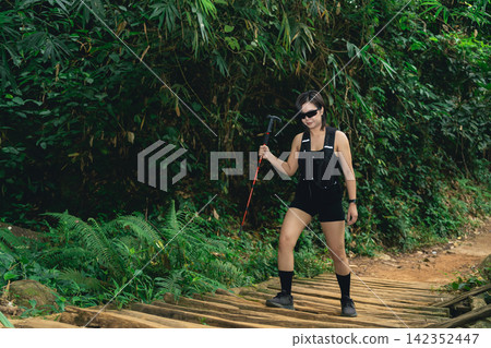 Woman with black outfit and sunglasses hiking on wooden path surrounded by lush green forest vegetation in daytime Woman with black outfit and sunglasses hiking on wooden path surrounded by lush green forest vegetation in daytime 142352447