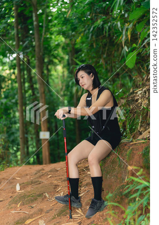 Young woman resting on dirt trail in dense green forest while hiking with trekking pole and athletic wear Young woman resting on dirt trail in dense green forest while hiking with trekking pole and athletic wear 142352572