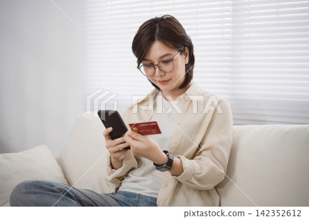 Woman Shopping Online on Smartphone While Sitting on Couch in Bright Living Room During Daylight Woman Shopping Online on Smartphone While Sitting on Couch in Bright Living Room During Daylight 142352612