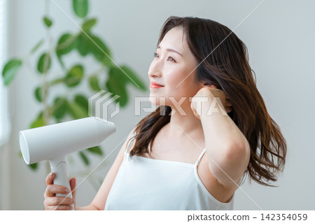 一位 40 多歲的女性正在用吹風機吹乾頭髮：頭髮護理和護髮習慣。 142354059