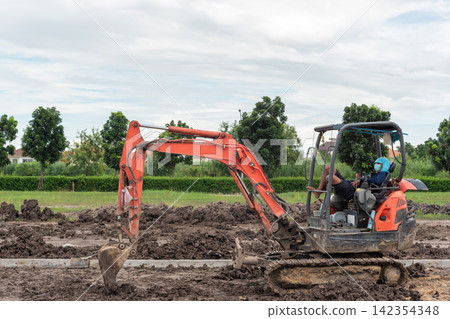 Construction worker operating compact excavator to dig move soil muddy outdoor worksite during land development earthwork Construction worker operating compact excavator to dig move soil muddy outdoor worksite during land development earthwork 142354348