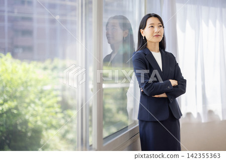 一位年輕的日本女商人雙手交叉，站在明亮的窗戶旁。 142355363