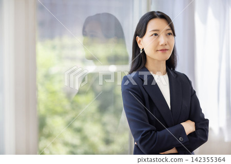 一位年輕的日本女商人雙手交叉，站在明亮的窗戶旁。 142355364
