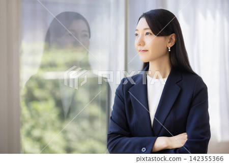 A young Japanese businesswoman stands with her arms crossed by a bright window. A young Japanese businesswoman stands with her arms crossed by a bright window. 142355366