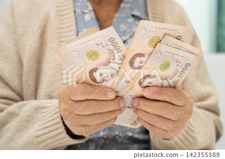 Asian elderly woman holding Thai banknotes money in hand. Poverty, saving problem in retirement. Asian elderly woman holding Thai banknotes money in hand. Poverty, saving problem in retirement. 142355389
