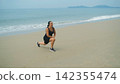 Young woman in black sportswear performing a stretching exercise with one leg extended on a sandy beach with calm sea behind. Flexibility training concept 142355474