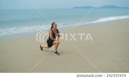 Young woman in black sportswear performing a stretching exercise with one leg extended on a sandy beach with calm sea behind. Flexibility training concept Young woman in black sportswear performing a stretching exercise with one leg extended on a sandy beach with calm sea behind. Flexibility training concept 142355474