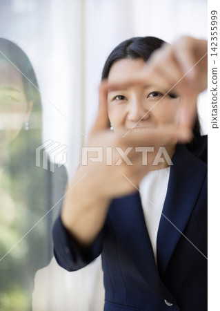 A young Japanese businesswoman makes a frame with her fingers and smiles while looking at the camera. A young Japanese businesswoman makes a frame with her fingers and smiles while looking at the camera. 142355999