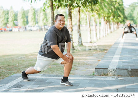 overweight man smiling while stretching thigh muscles before exercising in park overweight man smiling while stretching thigh muscles before exercising in park 142356174