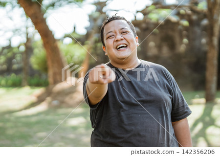 fat man smiles with a pointing gesture in the outdoor background 142356206
