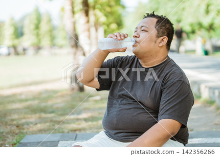 overweight man sitting drinking water with a bottle after a tired exercise overweight man sitting drinking water with a bottle after a tired exercise 142356266