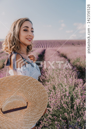 Woman lavender field smiling while holding a large straw hat in a blooming purple flower garden under blue sky Woman lavender field smiling while holding a large straw hat in a blooming purple flower garden under blue sky 142356526