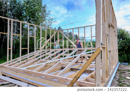Wall raising shows two builders manually lifting completed wall frame into place as timber frame house takes shape during construction. Wall raising shows two builders manually lifting completed wall frame into place as timber frame house takes shape during construction. 142357222