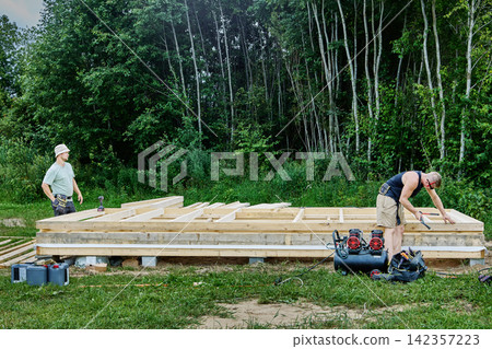 Construction of timber frame house on foundation of concrete blocks, workers assemble wooden floor frame during building construction. Construction of timber frame house on foundation of concrete blocks, workers assemble wooden floor frame during building construction. 142357223