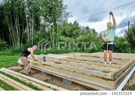 Foundation on concrete blocks supports timber frame construction, workers assemble wooden floor structure during building process. 142357224