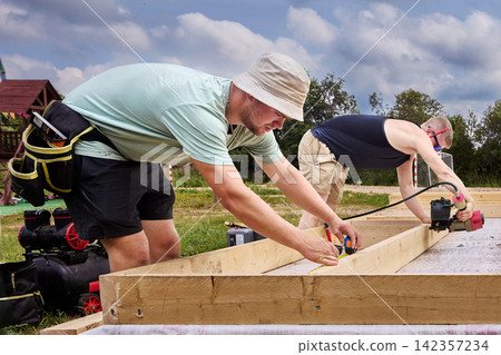 Construction workers assemble timber floor frame, one measuring wooden beam while another uses pneumatic nail gun to secure boards. Construction workers assemble timber floor frame, one measuring wooden beam while another uses pneumatic nail gun to secure boards. 142357234