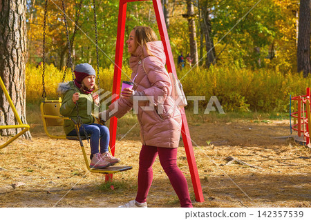Mother in pink jacket shakes her child daughter on swing in sunny autumn park Mother in pink jacket shakes her child daughter on swing in sunny autumn park 142357539
