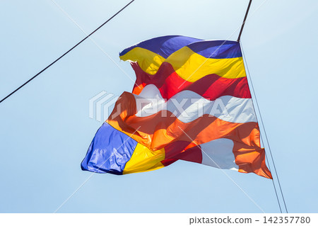 Colorful Buddhist flag with vertical stripes waving on a rope against clear blue sky Colorful Buddhist flag with vertical stripes waving on a rope against clear blue sky 142357780