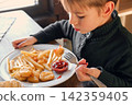 Child Eating French Fries with Tomato Sauce in Restaurant Indoors. Little Kid with Blond Hair Enjoy Unhealthy Fast Food. lunch. Caucasian Boy Eats Potato Chips at the Table. Six Years Old Toddler 142359405