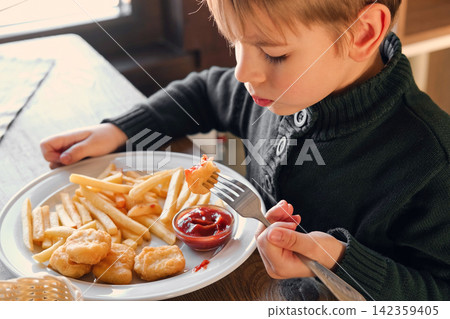 Child Eating French Fries with Tomato Sauce in Restaurant Indoors. Little Kid with Blond Hair Enjoy Unhealthy Fast Food. lunch. Caucasian Boy Eats Potato Chips at the Table. Six Years Old Toddler 142359405