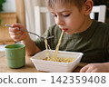 6 years old boy eats instant noodle from box. Child is eating spaghetti. Lunch time. A good appetite. Tasty children food. Close-up portrait. Carbs dinner. Fast food. Kid eating by himself with fork. 142359408