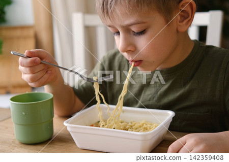 6 years old boy eats instant noodle from box. Child is eating spaghetti. Lunch time. A good appetite. Tasty children food. Close-up portrait. Carbs dinner. Fast food. Kid eating by himself with fork. 142359408