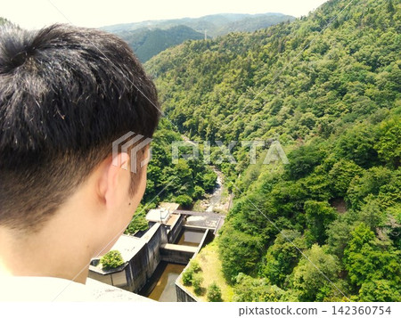 一個男人的背影,他俯瞰著鬱鬱蔥蔥的山谷和水壩。 一個男人的背影,他俯瞰著鬱鬱蔥蔥的山谷和水壩。 142360754