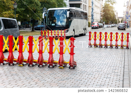 Expandable red and yellow security barriers block cobblestone street while coach waits behind temporary checkpoint. Urban traffic control creates protected route during public event in Magdeburg 142360832
