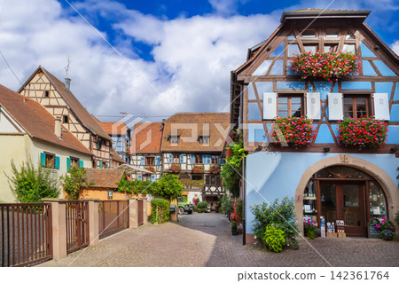 Picturesque historic street in Eguisheim Alsace France. Traditional half timbered architecture and facade under cloudy sky Picturesque historic street in Eguisheim Alsace France. Traditional half timbered architecture and facade under cloudy sky 142361764