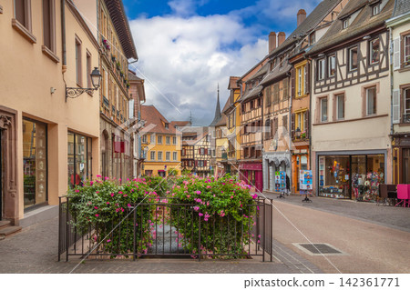View of a historic street in Colmar Alsace France with floral decoration. Traditional architecture under partly cloudy sky View of a historic street in Colmar Alsace France with floral decoration. Traditional architecture under partly cloudy sky 142361771