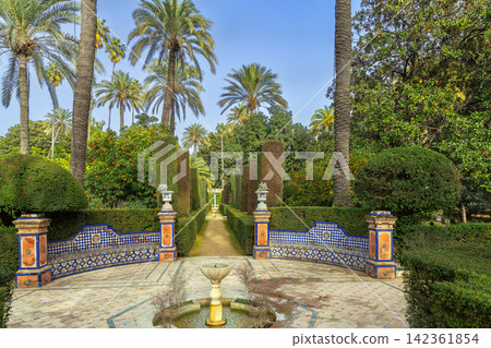 Decorative alley in Alcazar gardens of Seville Spain with small fountain. Ceramic tile benches under clear blue sky 142361854