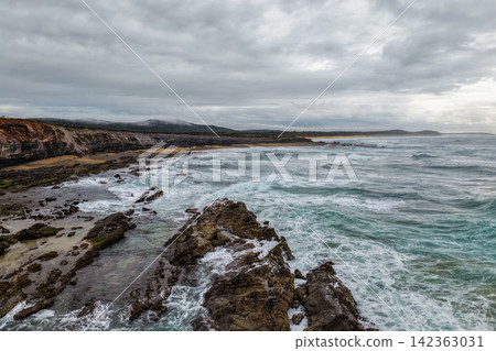 Aerial view of ocean waves crashing onto the rugged rocky coastline at Lake Arragan Beach beneath a cloudy sky, showcasing dramatic surf, exposed rock formations and the wild Pacific coastline Aerial view of ocean waves crashing onto the rugged rocky coastline at Lake Arragan Beach beneath a cloudy sky, showcasing dramatic surf, exposed rock formations and the wild Pacific coastline 142363031
