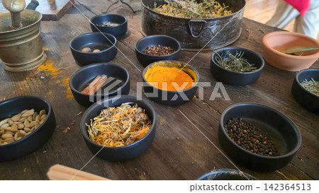 Assorted spices and dried herbs are arranged in bowls on a rustic wooden worktable during traditional food preparation. Concepts: culinary craftsmanship, natural ingredients, cultural tradition. Assorted spices and dried herbs are arranged in bowls on a rustic wooden worktable during traditional food preparation. Concepts: culinary craftsmanship, natural ingredients, cultural tradition. 142364513