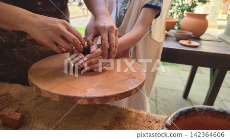 Adult hands guide a child while shaping clay on a pottery wheel during a ceramics workshop. Concepts: education, creativity, craftsmanship, learning, family, mentoring, tradition, handmade Adult hands guide a child while shaping clay on a pottery wheel during a ceramics workshop. Concepts: education, creativity, craftsmanship, learning, family, mentoring, tradition, handmade 142364606