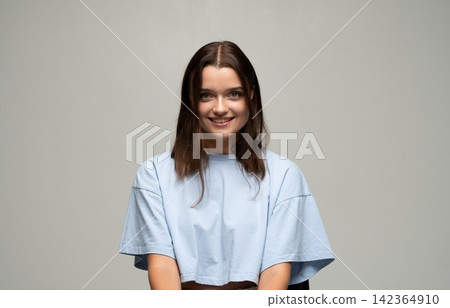 Young woman with brown hair smiling, posing in light blue t-shirt, embracing happiness and confidence in studio portrait 142364910
