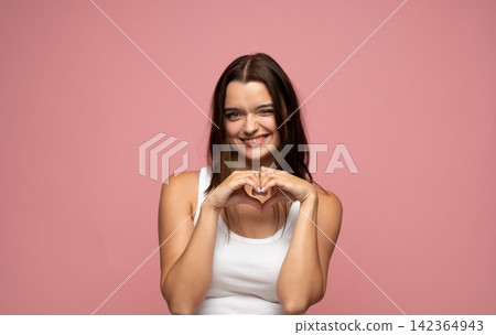 Young woman making heart shape gesture with hands and winking, expressing love and happiness on pink background 142364943