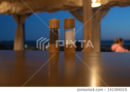 Two wooden salt and pepper grinders standing on a restaurant table at dusk, creating a warm dining atmosphere with soft bokeh, ambient lighting and an elegant outdoor cafe setting Two wooden salt and pepper grinders standing on a restaurant table at dusk, creating a warm dining atmosphere with soft bokeh, ambient lighting and an elegant outdoor cafe setting 142366028