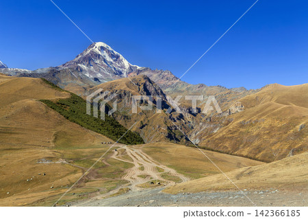 Majestic Mount Kazbek rises behind Gergeti Trinity Church in Georgia. High angle view in bright autumn afternoon light Majestic Mount Kazbek rises behind Gergeti Trinity Church in Georgia. High angle view in bright autumn afternoon light 142366185