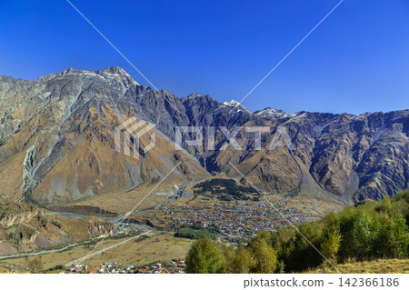 High angle view of Stepantsminda town in the Caucasus mountains, Georgia. Scenic panorama from a hill in sunny daylight High angle view of Stepantsminda town in the Caucasus mountains, Georgia. Scenic panorama from a hill in sunny daylight 142366186