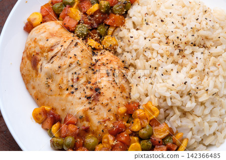 a dish of homemade roasted chicken breast and dice carrots, beans, corns with brown rice on dark red wooden table and cutlery 142366485