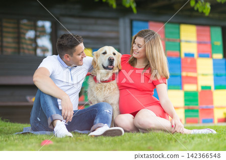 Happy young pregnant couple sitting on the grass in a park, smiling lovingly at their Golden Retriever dog positioned between them in front of a colorful backdrop Happy young pregnant couple sitting on the grass in a park, smiling lovingly at their Golden Retriever dog positioned between them in front of a colorful backdrop 142366548