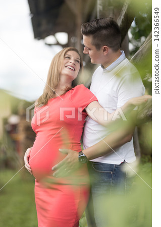 Three-quarter vertical portrait of a joyful pregnant woman in a red dress leaning back against her husband as they both hold her baby bump outdoors in nature Three-quarter vertical portrait of a joyful pregnant woman in a red dress leaning back against her husband as they both hold her baby bump outdoors in nature 142366549