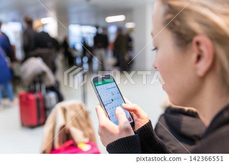 Close-up over-the-shoulder view of a female traveler checking a calendar app on her smartphone while sitting in a busy airport terminal near luggage 142366551